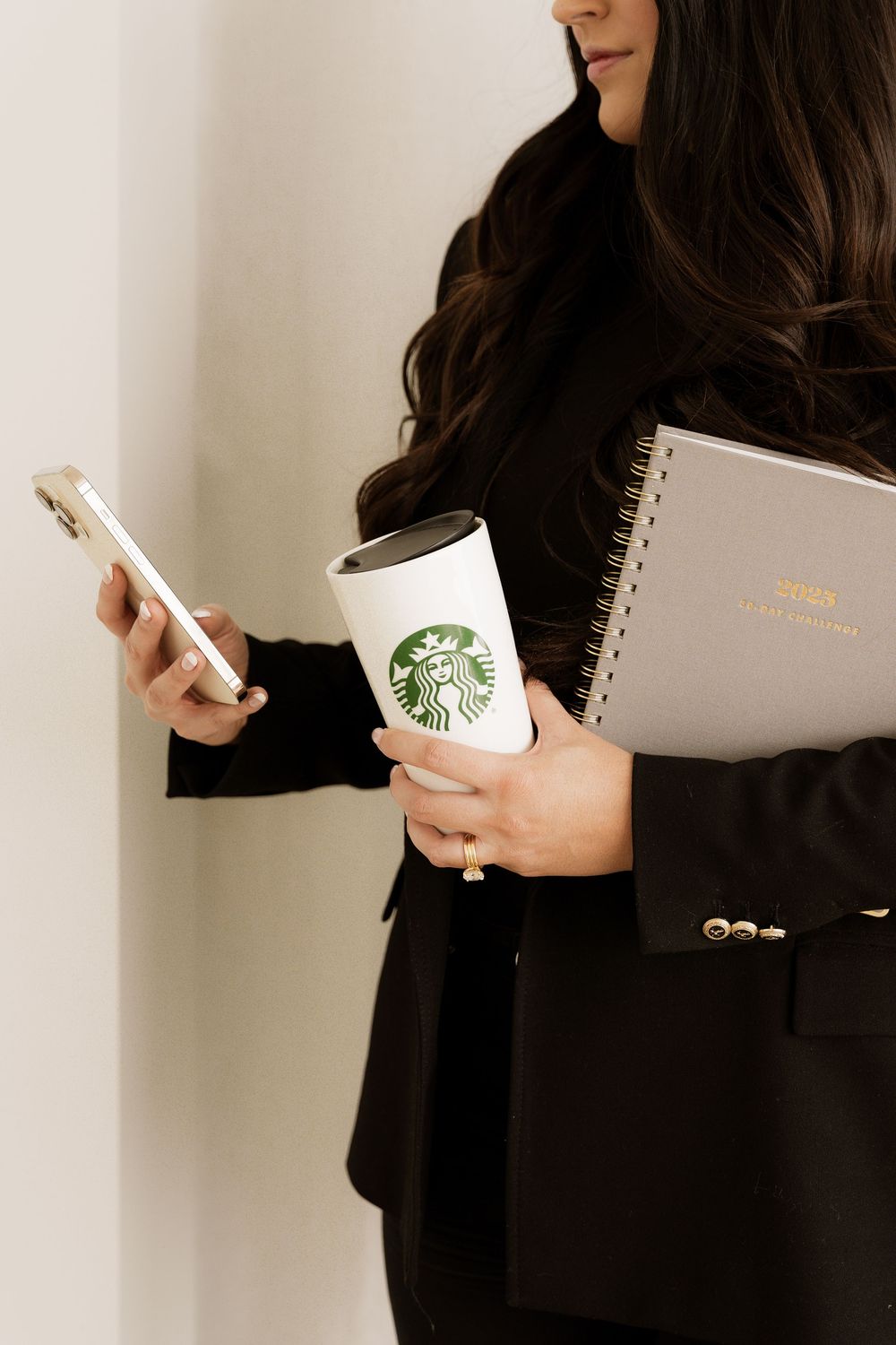 Professional woman in black blazer holding Starbucks cup, phone and notebook while working on the go.