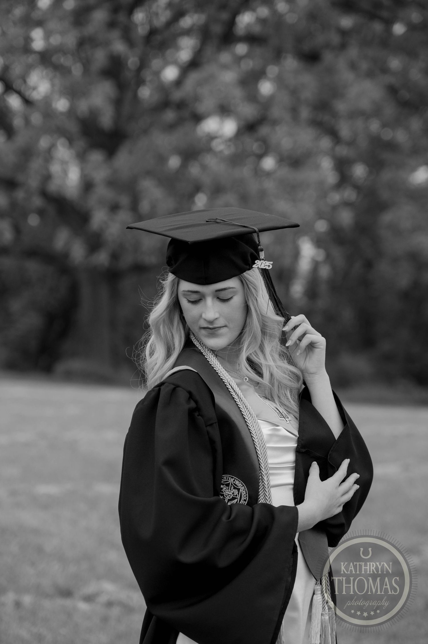Graduate in cap and gown stands in a green outdoor setting while adjusting their graduation attire.