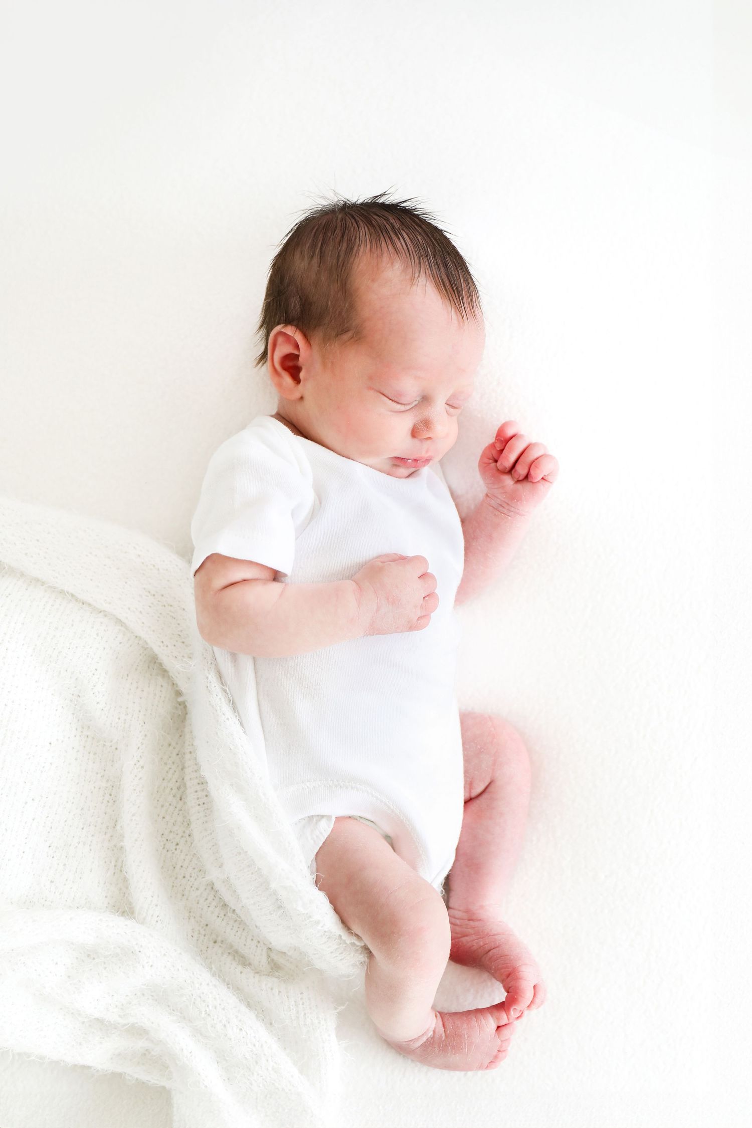 Sleeping newborn baby wearing white onesie lying on soft white blanket in minimalist setting, during Newborn shoot in Nottingham