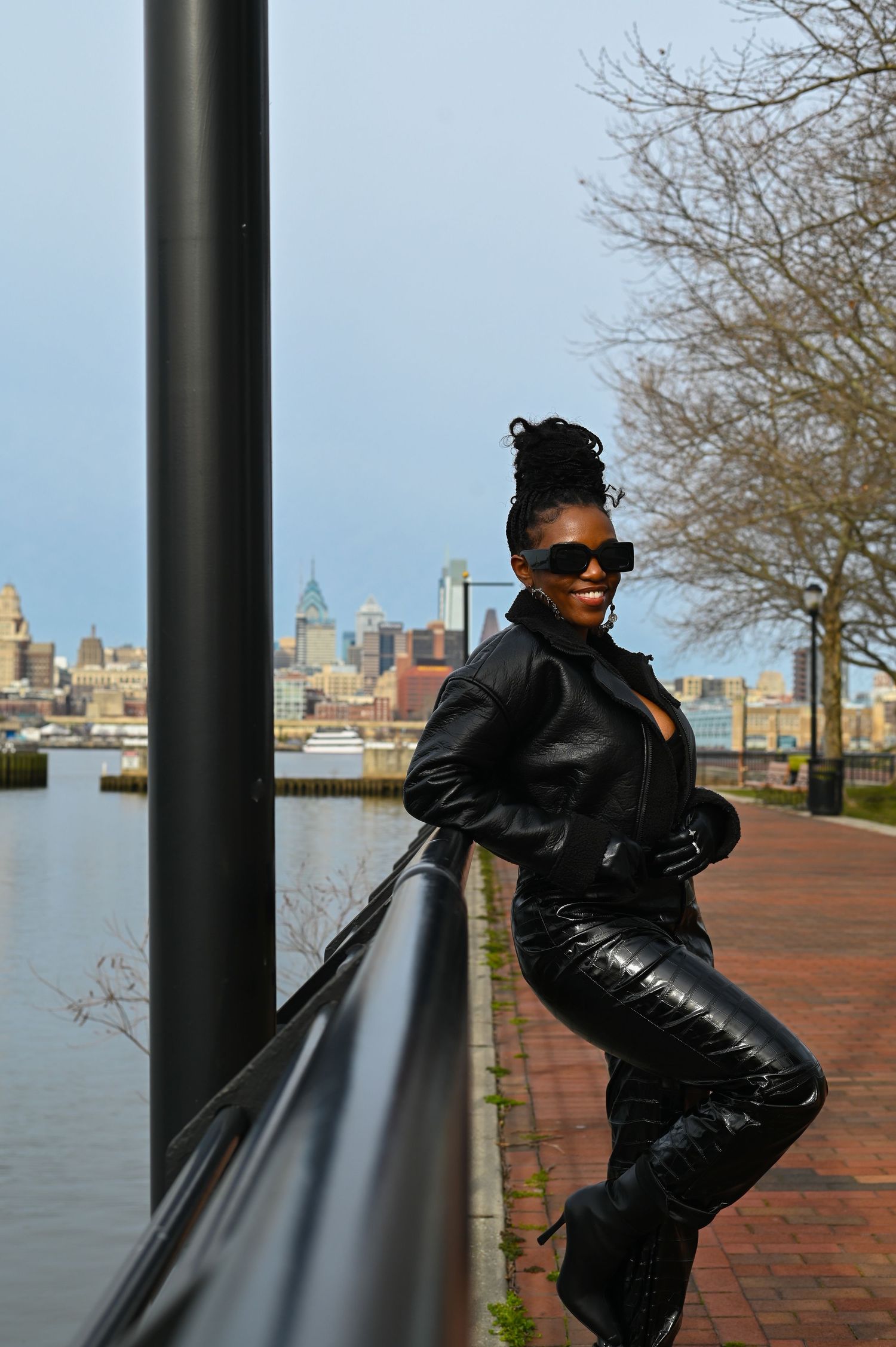 A fashionable person in an all-black outfit poses along a waterfront brick path with a city skyline in the background.