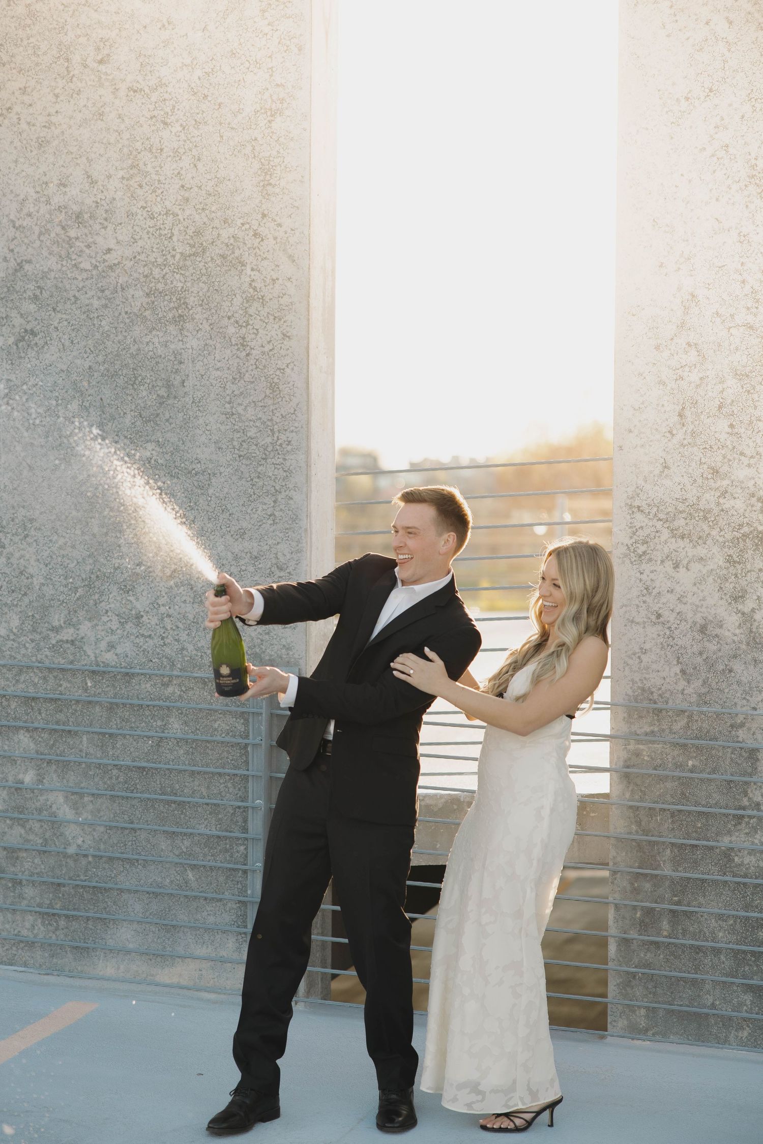 A couple in formal attire celebrate by popping champagne on a rooftop balcony during sunset.