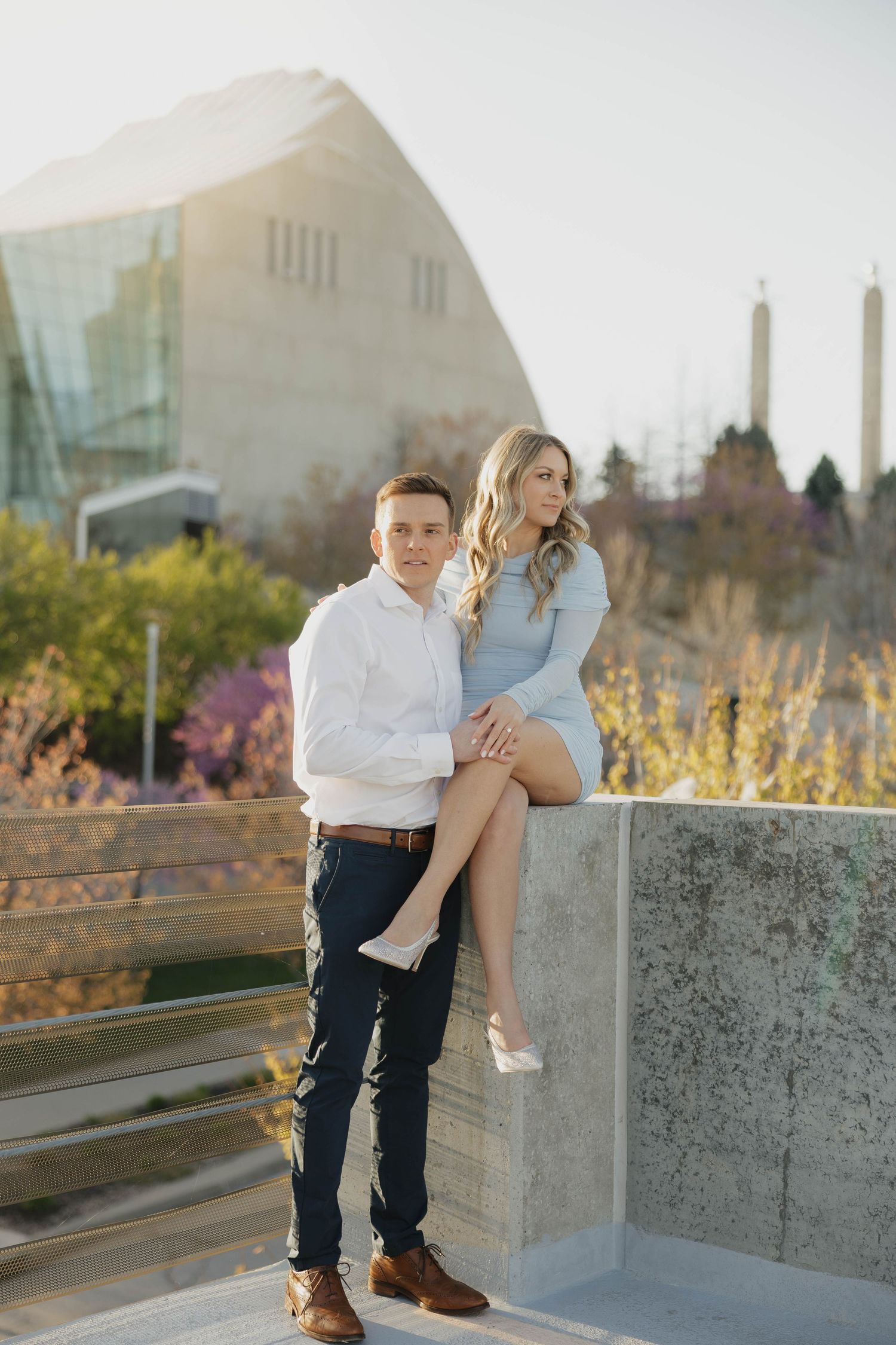 A couple poses together on an outdoor concrete ledge during a sunny autumn day with modern architecture in background.