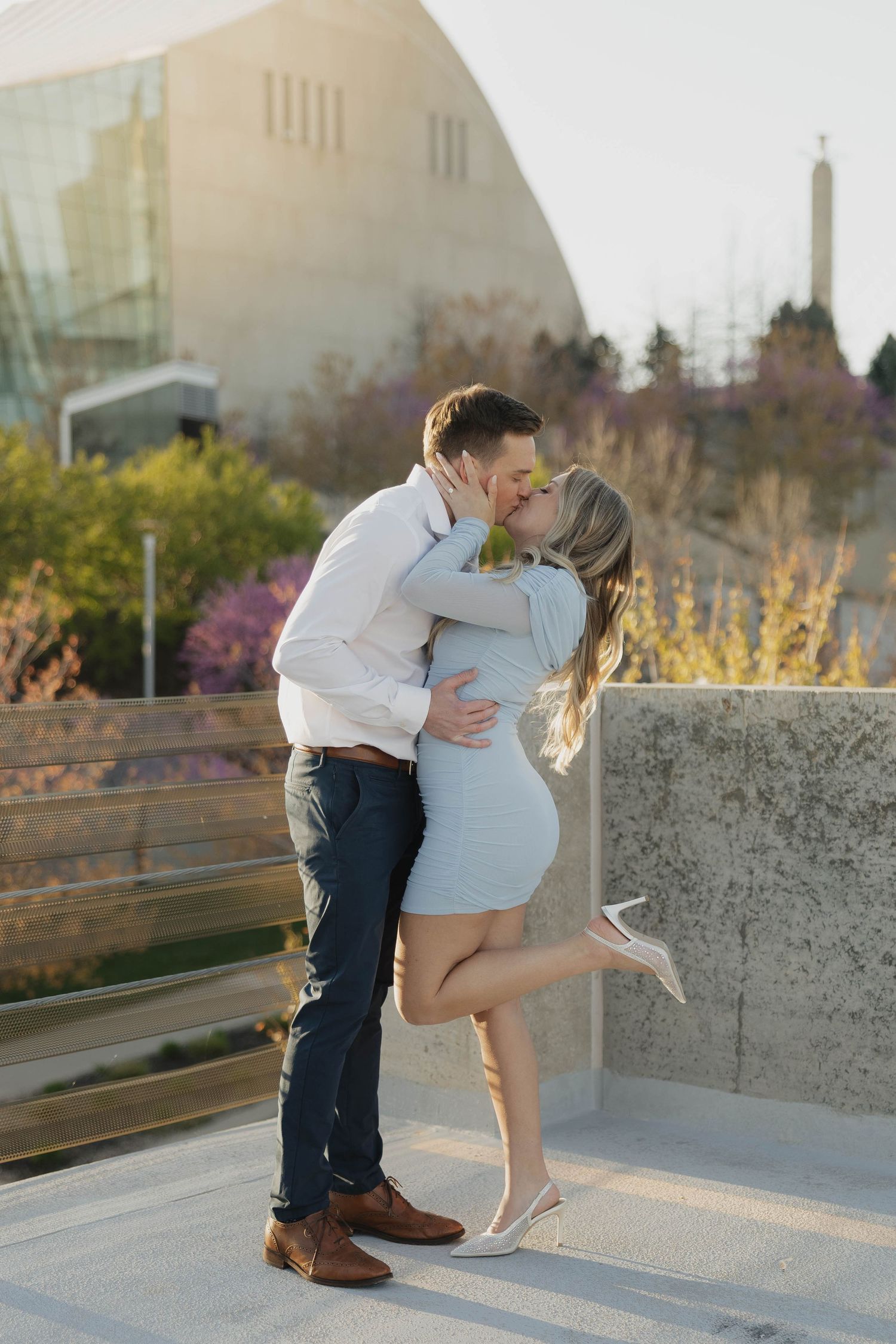 A couple shares a romantic kiss on a rooftop terrace during golden hour with blurred city buildings in the background.