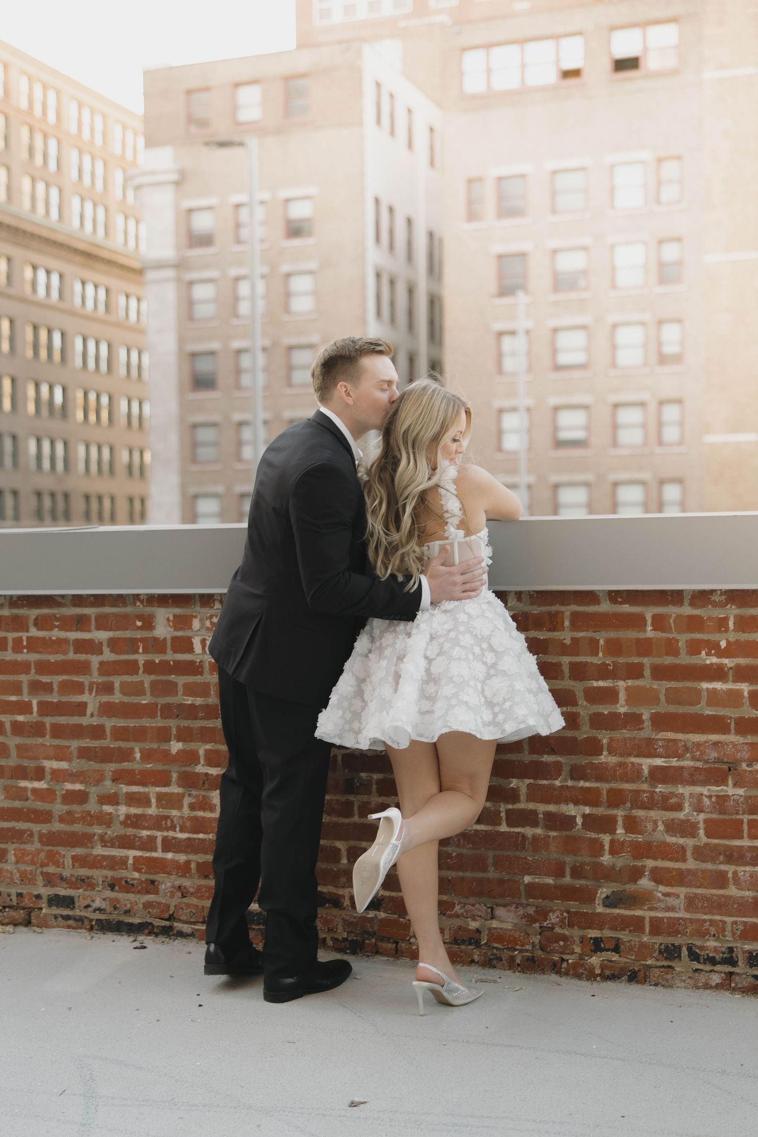A couple shares a romantic moment on a brick rooftop with apartment buildings in the background.