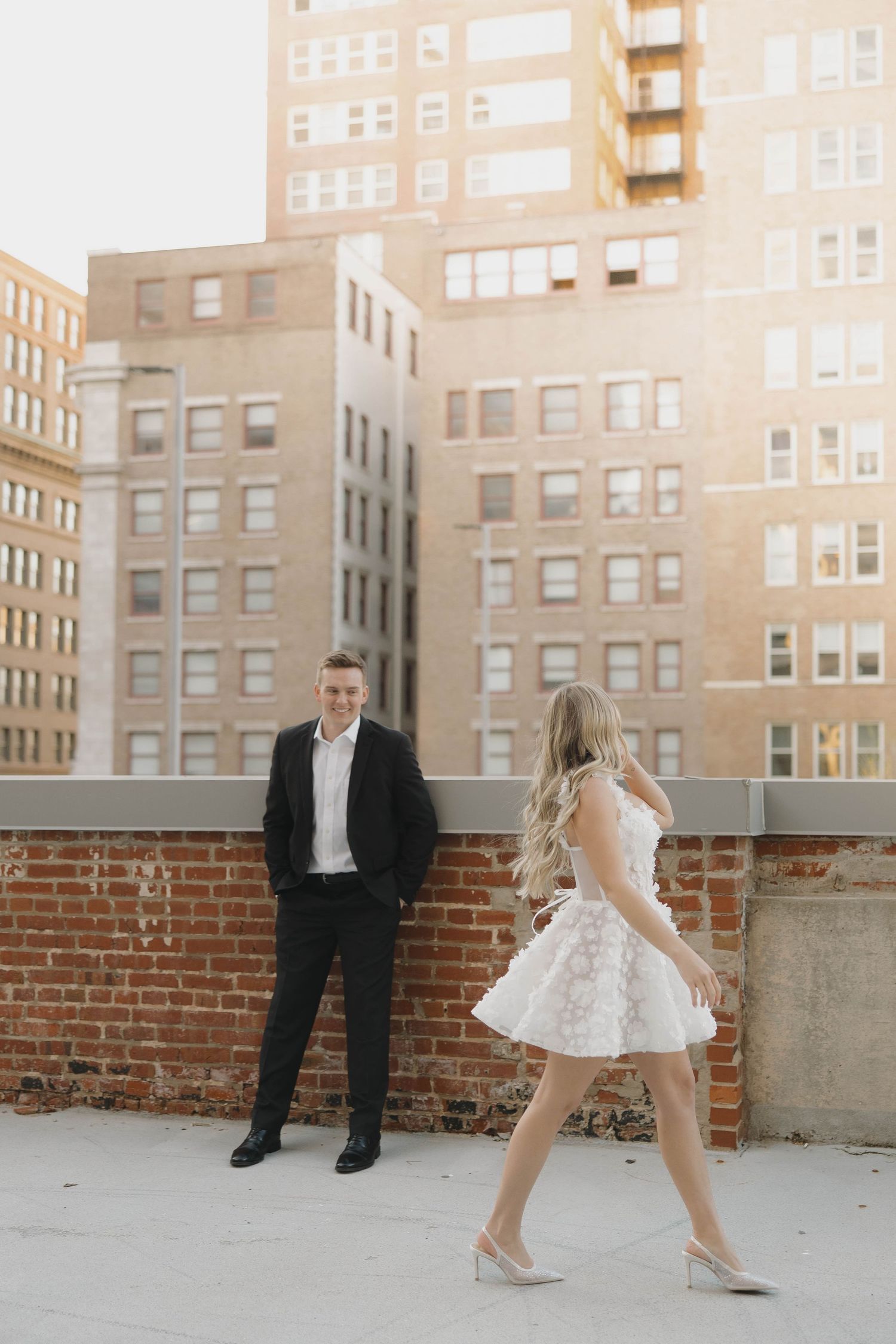 Elegant couple poses on urban rooftop with brick wall and apartment buildings in golden sunlight during photo session.