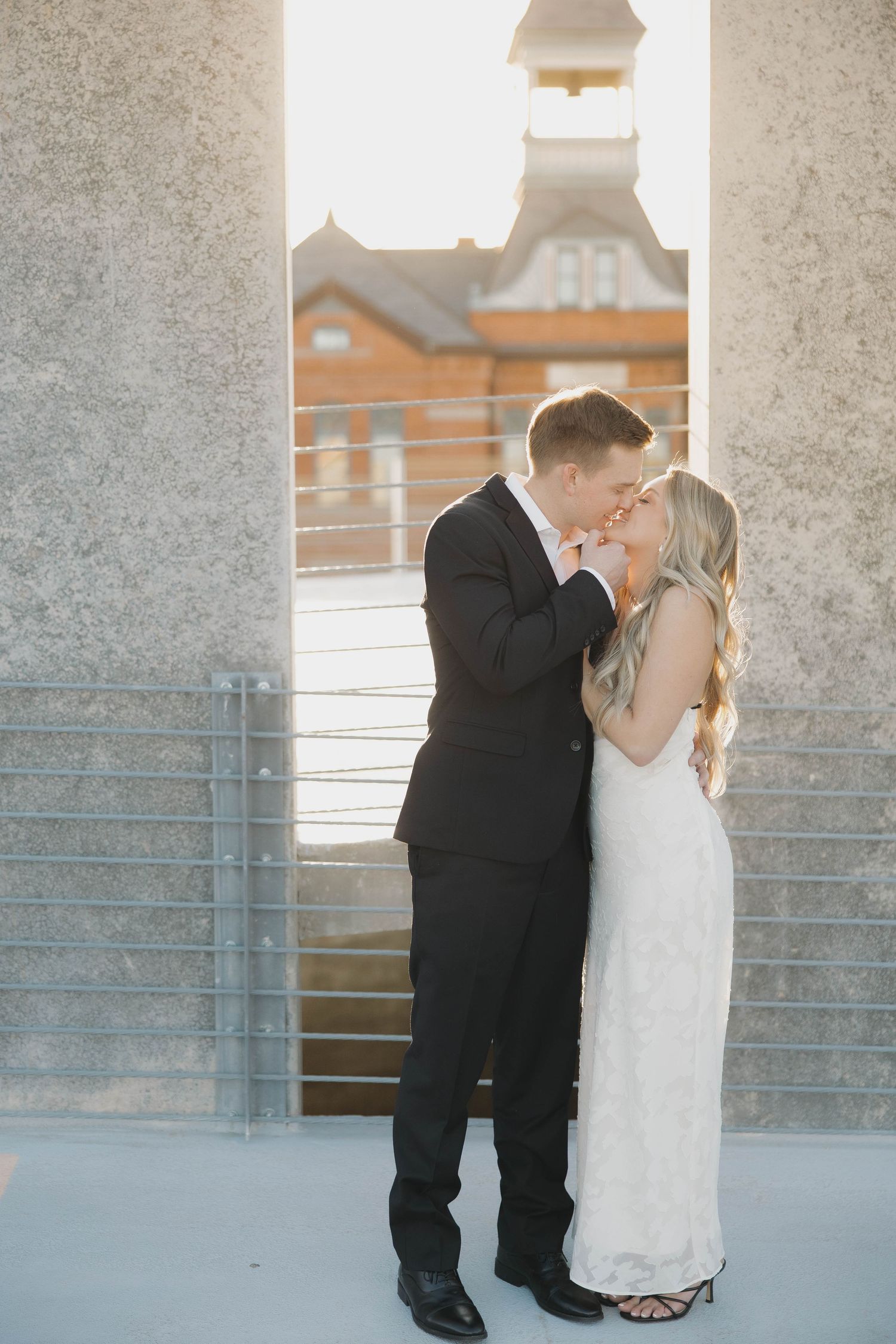Bride and groom share a romantic kiss against a sunlit backdrop of stone columns and modern architecture.