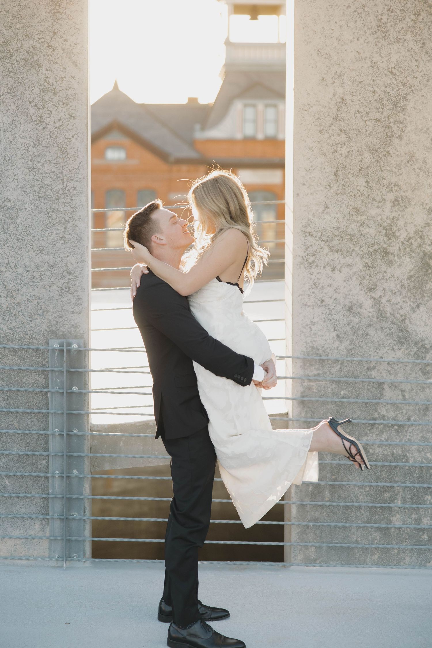 A romantic couple embraces in formal attire against a sunlit backdrop with architectural columns and metal railings.