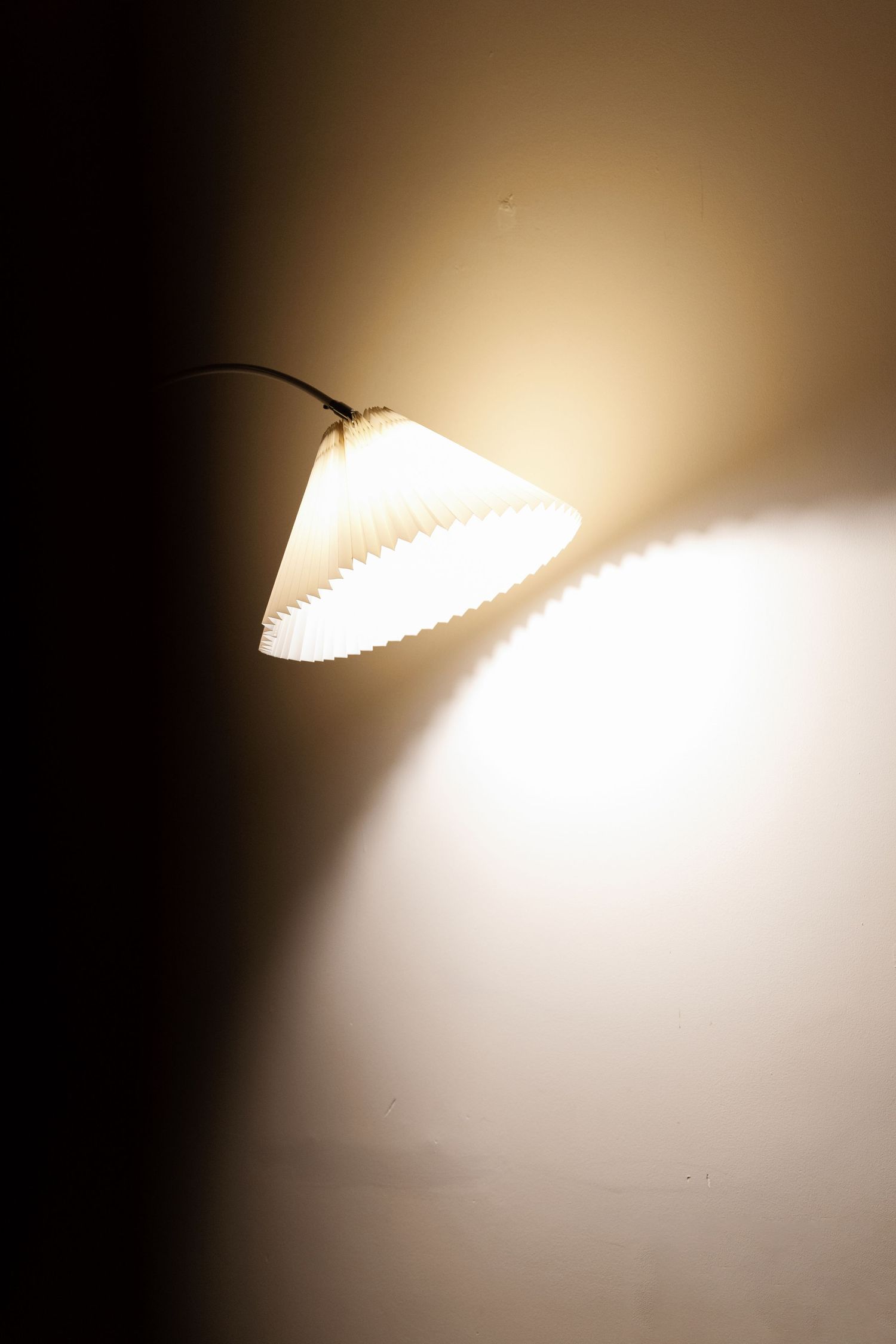 A glowing white pleated lamp shade casts warm light against a dark background creating dramatic shadows and contrast.