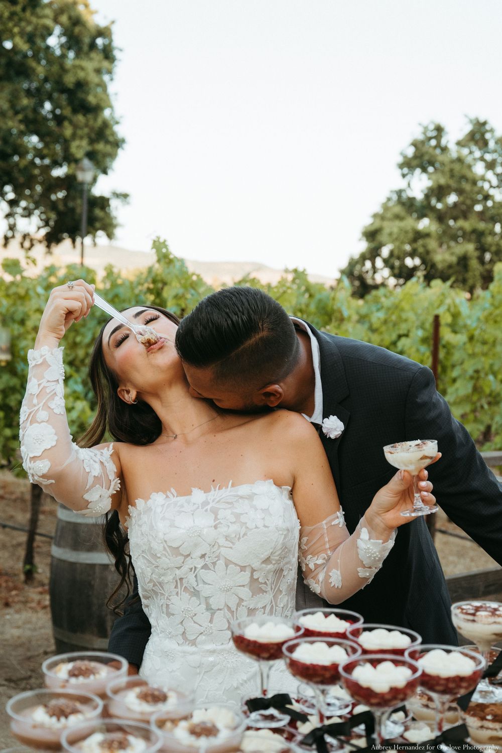 Couple celebrating at a wedding reception with champagne tower and decorative dessert display in an outdoor vineyard setting.