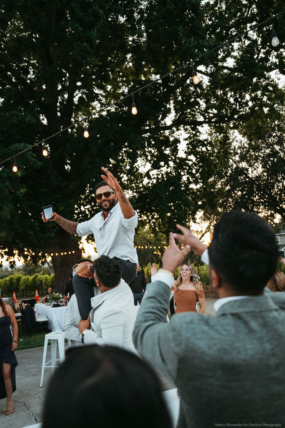 Guests celebrate at an outdoor party with string lights hanging in trees as someone dances on a chair at dusk.