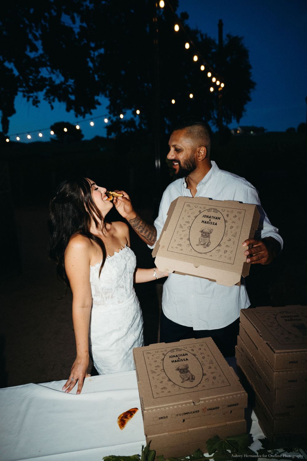 A couple holding pizza boxes shares a romantic nighttime moment under string lights at an outdoor event.