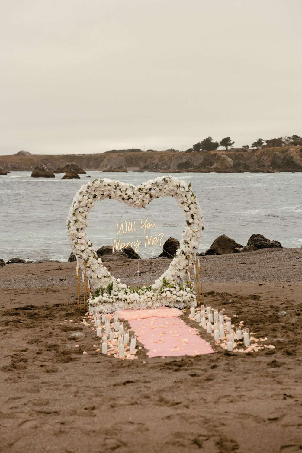 A heart-shaped floral arch on a rocky beach with a pink blanket set up for a romantic proposal.