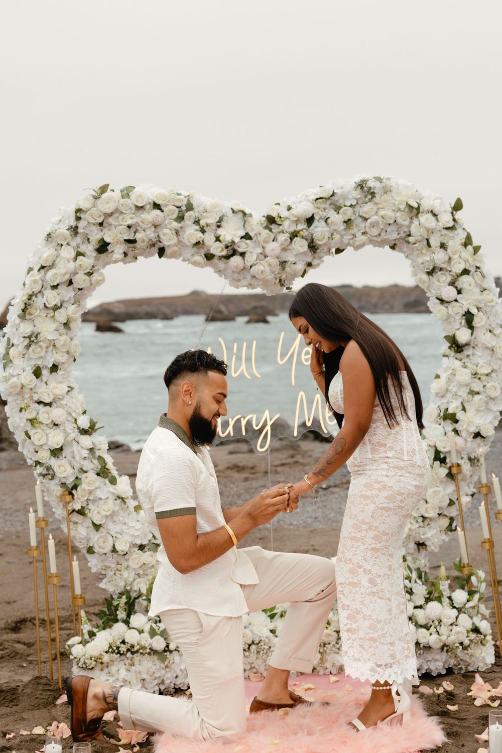 A romantic proposal scene on a beach with a beautiful white floral arch and pink rose petals scattered on the ground.