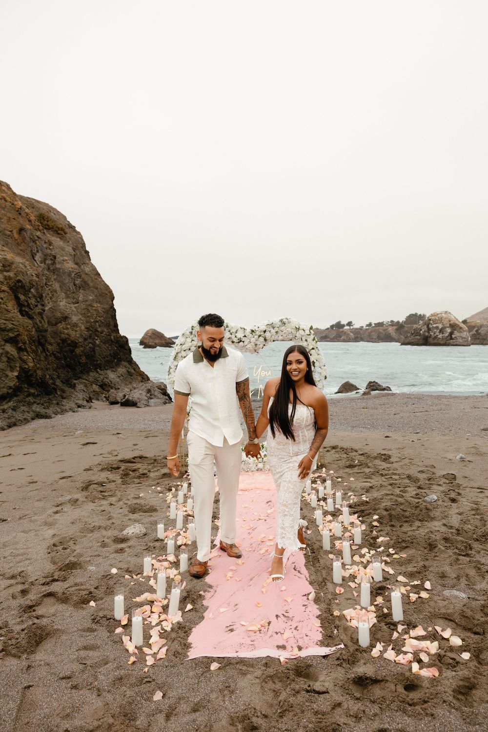 Two people create a heart shape with candles on a pink blanket at a rocky beach during golden hour.