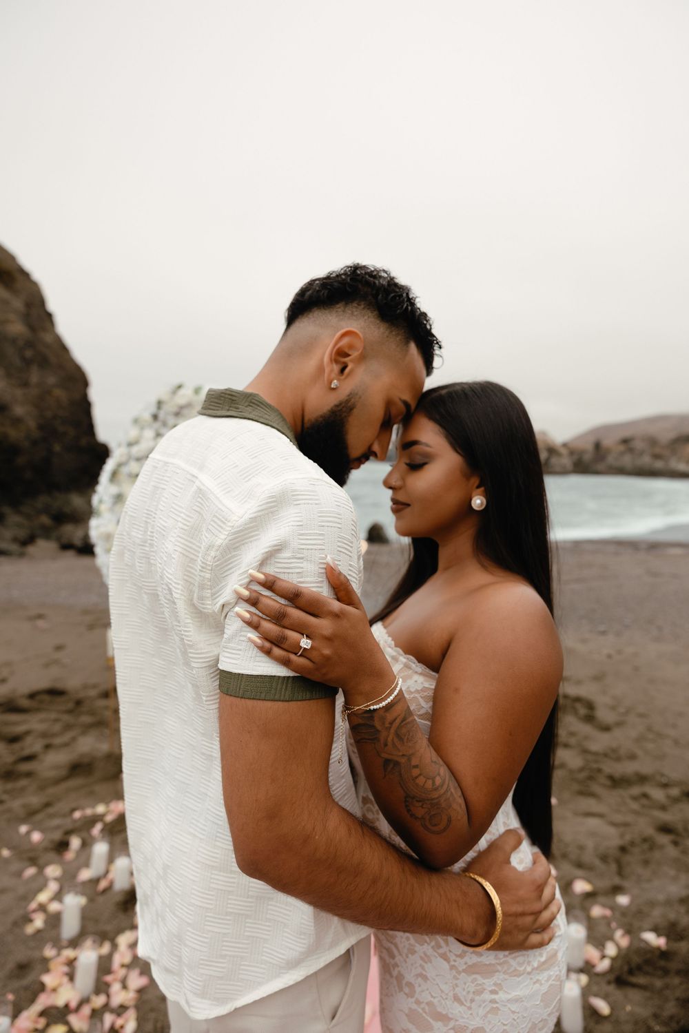 A romantic couple embraces on a moody beach in Bodega Bay, CA with rocks and overcast skies in the background.