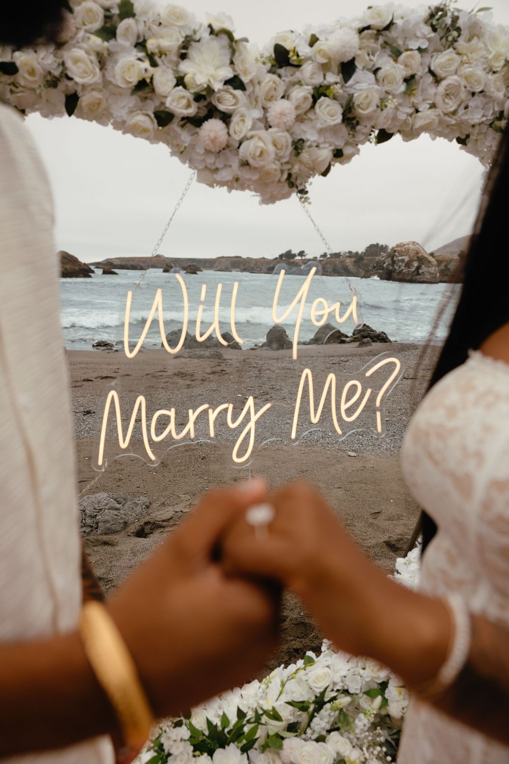 A romantic Bodega Bay, CA beach proposal setup with 'Will You Marry Me' written on a sign with the ocean behind and white flowers above.