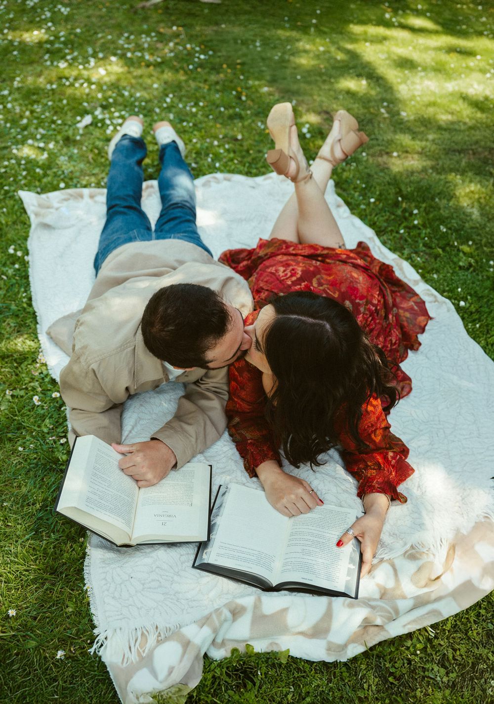 Two people lying together on a white blanket reading a book on grass in a garden setting.