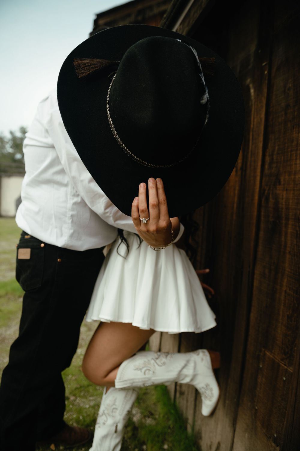 A romantic couple dressed in black and white embraces near a rustic wooden wall outdoors.