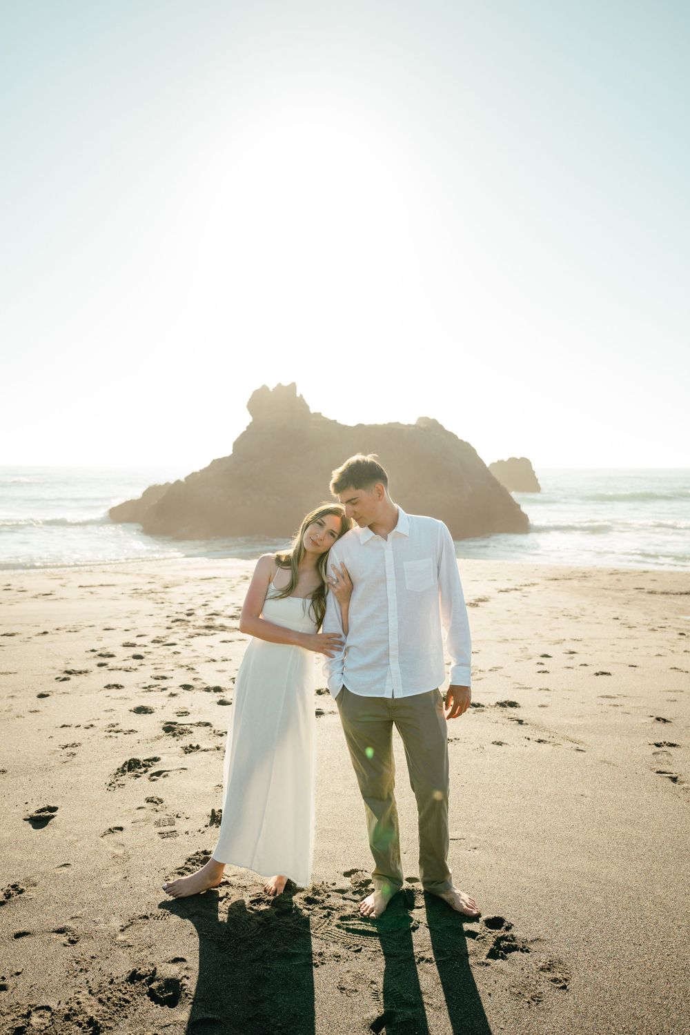 A couple in white and green attire share an intimate moment on a sunny beach with rocky cliffs in the background.