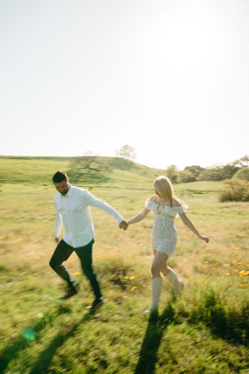 Couple holding hands running through sunny green meadow, woman in white dress and boots, man in white shirt and dark pants.