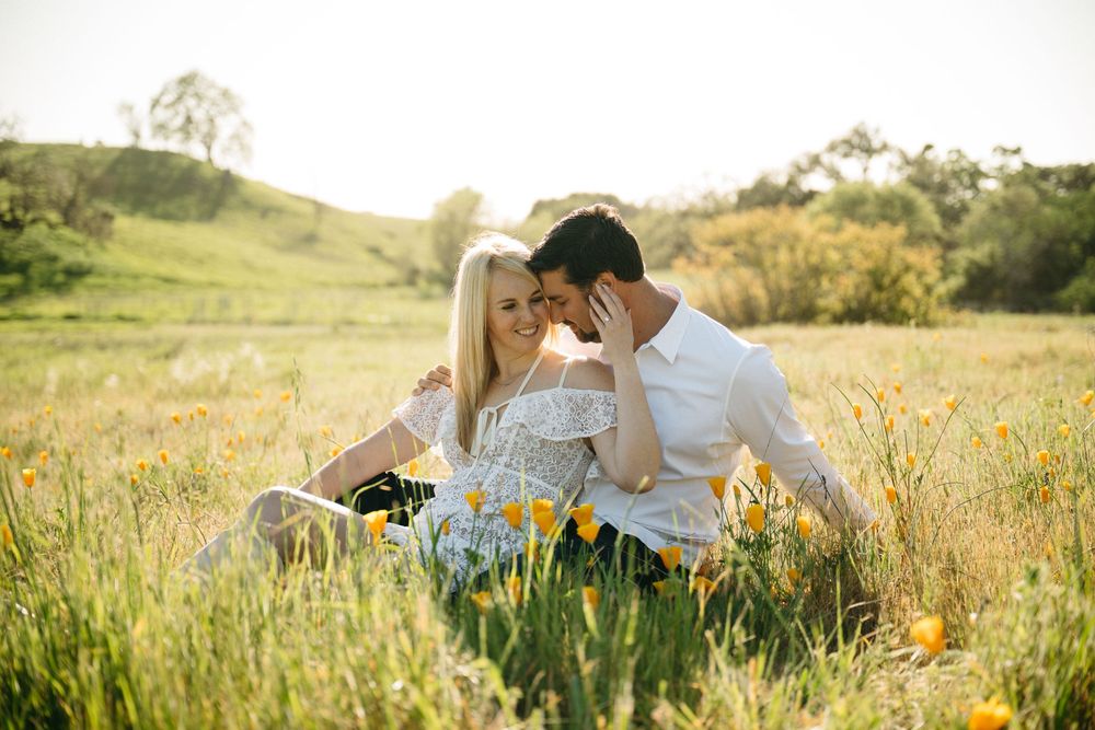 Couple sitting close together in a sunny golden field of wildflowers, sharing a romantic intimate moment outdoors.