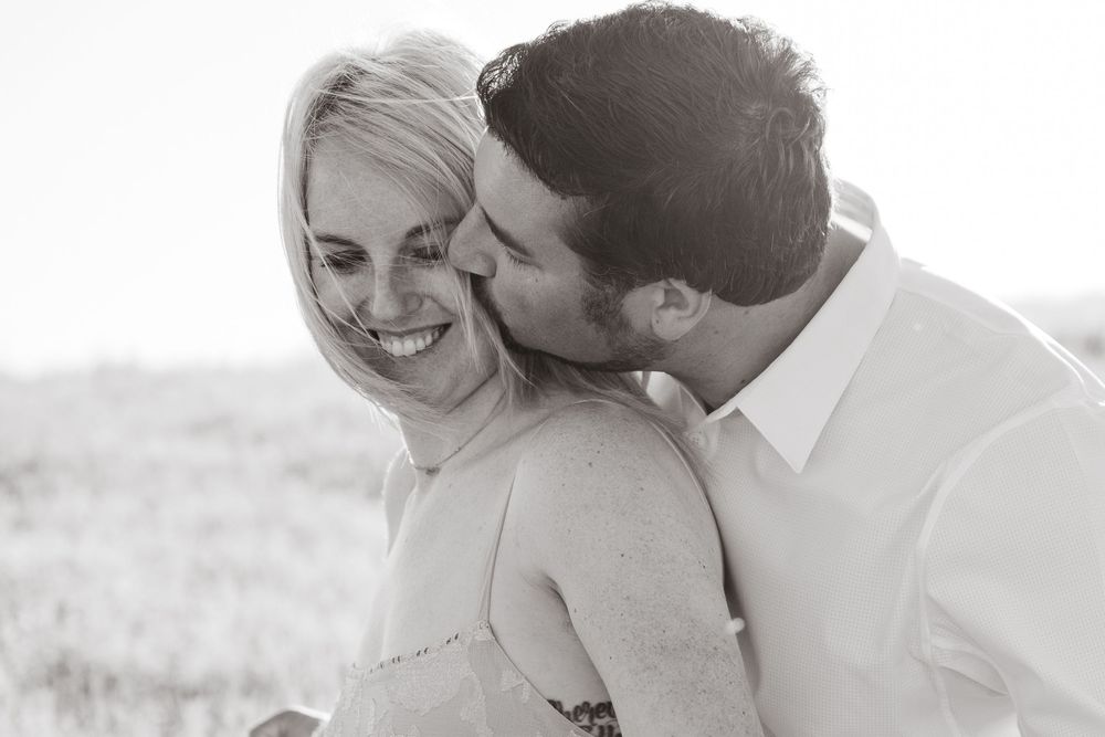 Black and white photo of a smiling woman being kissed on the cheek by a man outdoors.