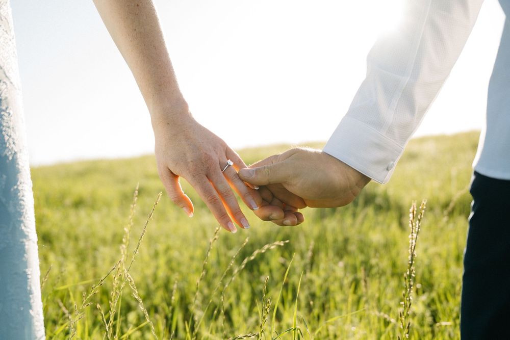 Couple holding hands in a green field, bride wearing engagement ring, romantic outdoor wedding photo in sunlit meadow.