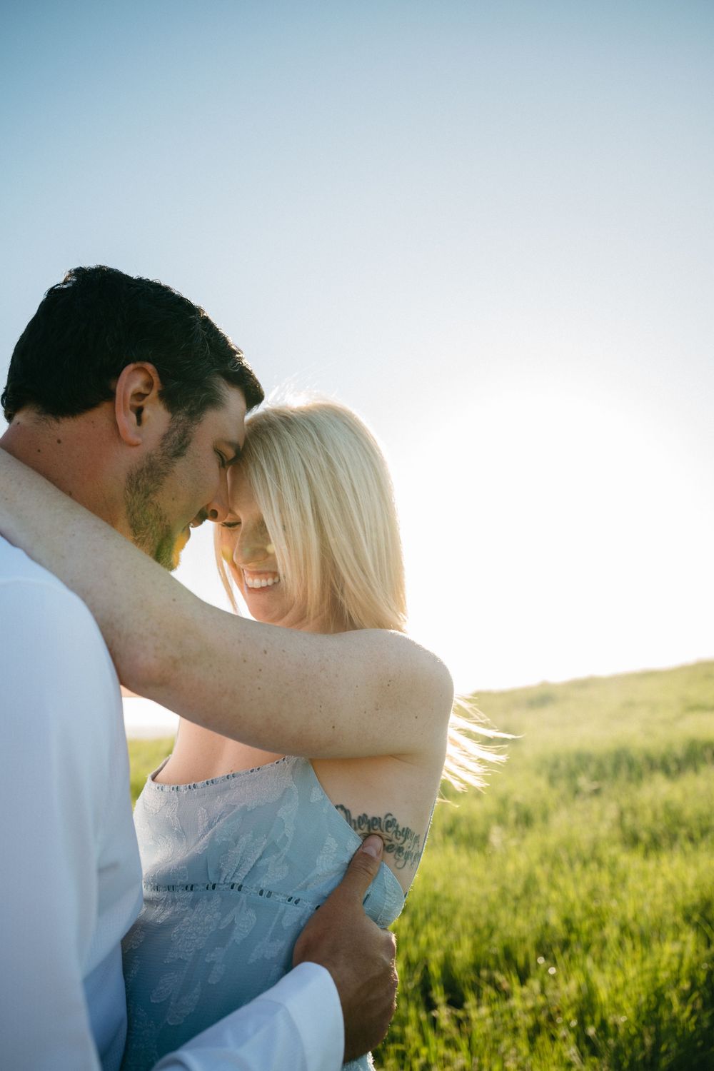 A joyful couple embraces in a sunlit green field, the smiling blonde woman in a blue dress nuzzling foreheads with her partner.