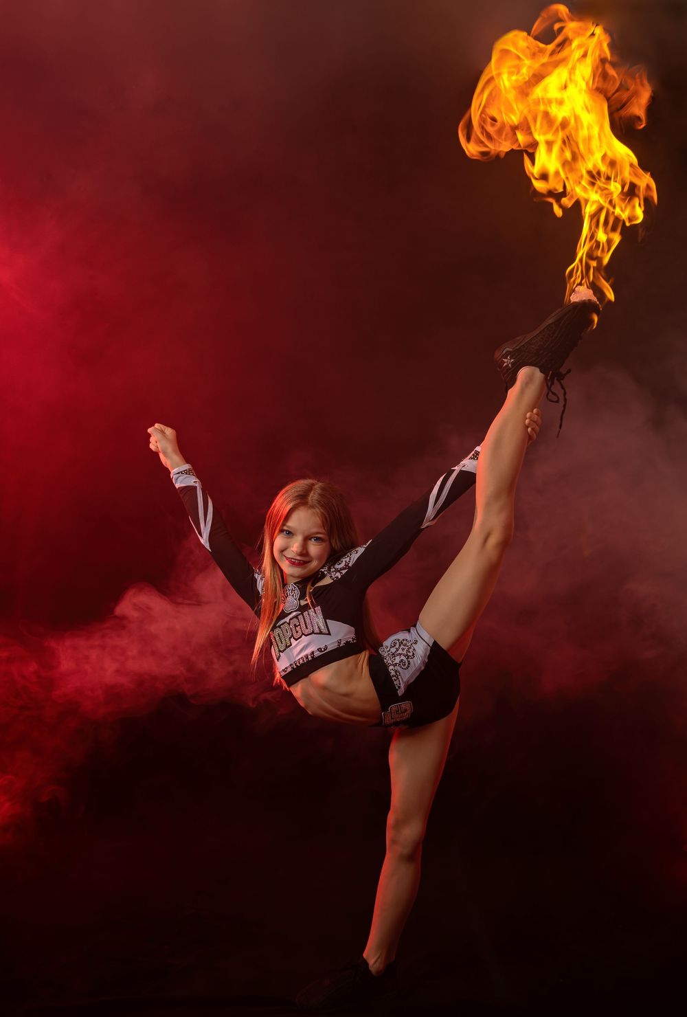 Young performer executing high kick with dramatic fire effect against red smoky background.