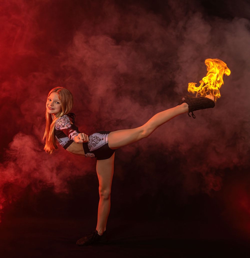 Dancer performs arabesque pose with fire prop in atmospheric red smoke setting.