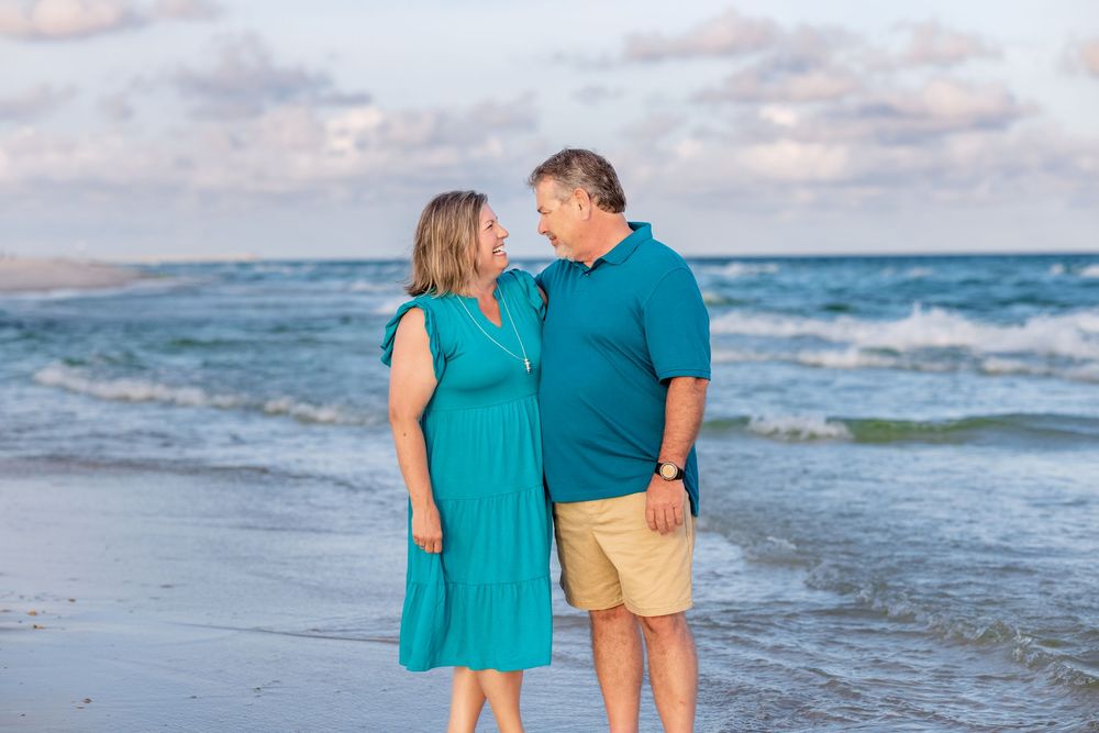 Two people in color-coordinated turquoise and yellow outfits stand together on a beach at sunset with ocean waves behind them.