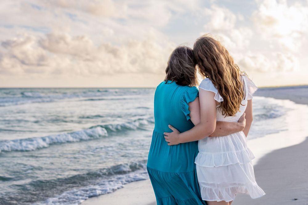Two people embrace while standing on a sandy beach at sunset with gentle ocean waves in the background.