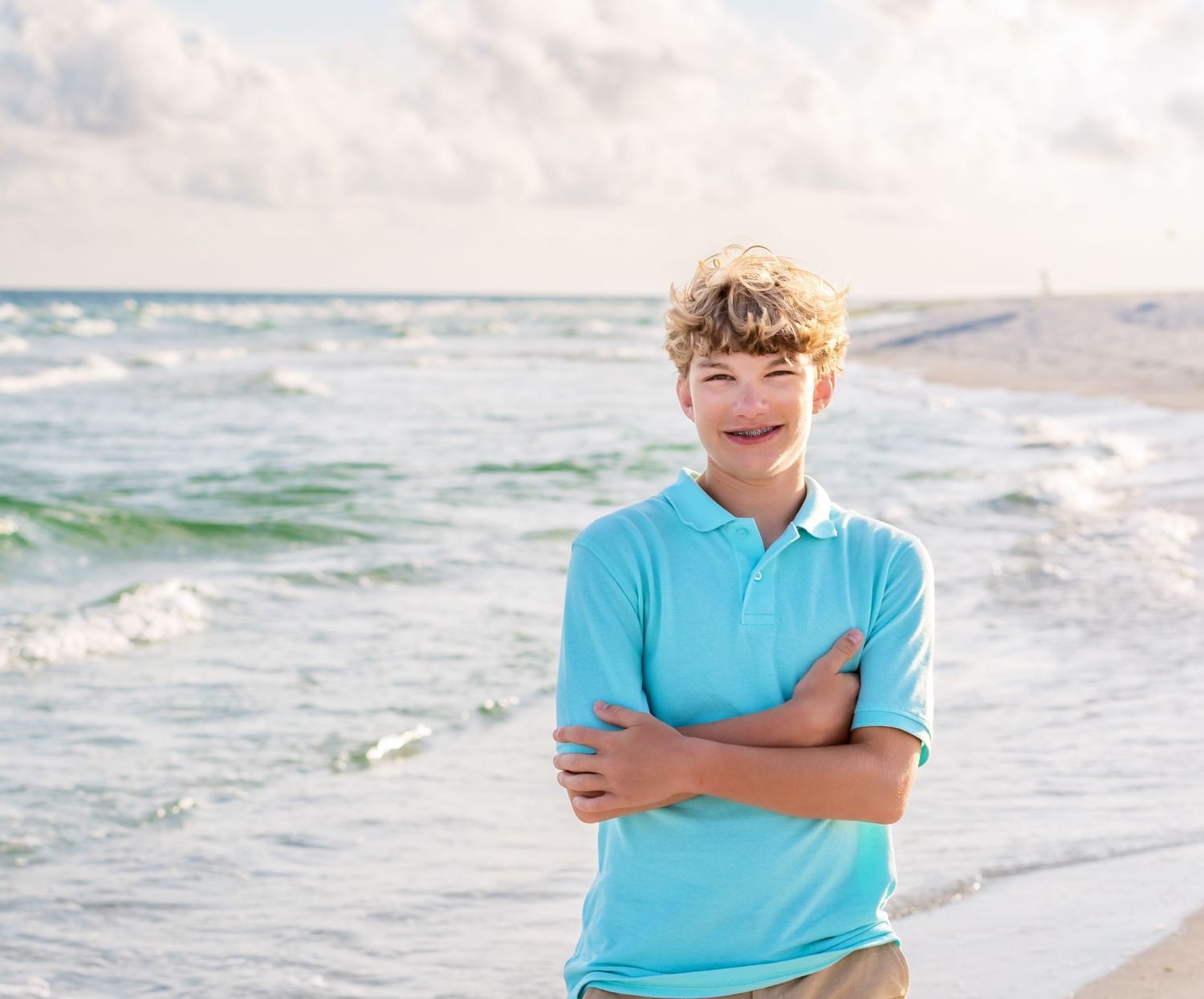 A beachgoer in a turquoise polo shirt standing with arms crossed along a scenic shoreline with crashing waves.