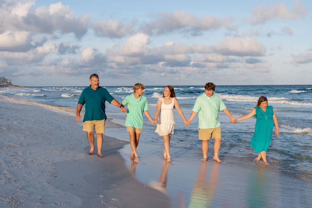 Family walking together along a scenic beach shoreline at sunset wearing coordinated turquoise and white outfits.
