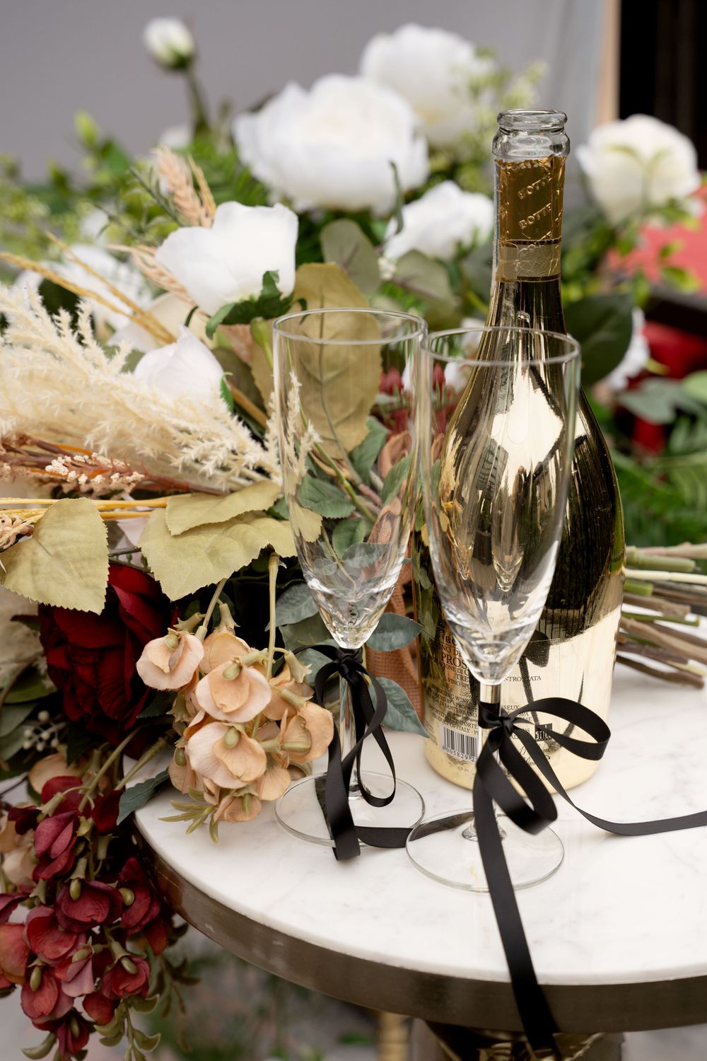 Champagne bottle and glasses surrounded by elegant floral arrangements on white table.