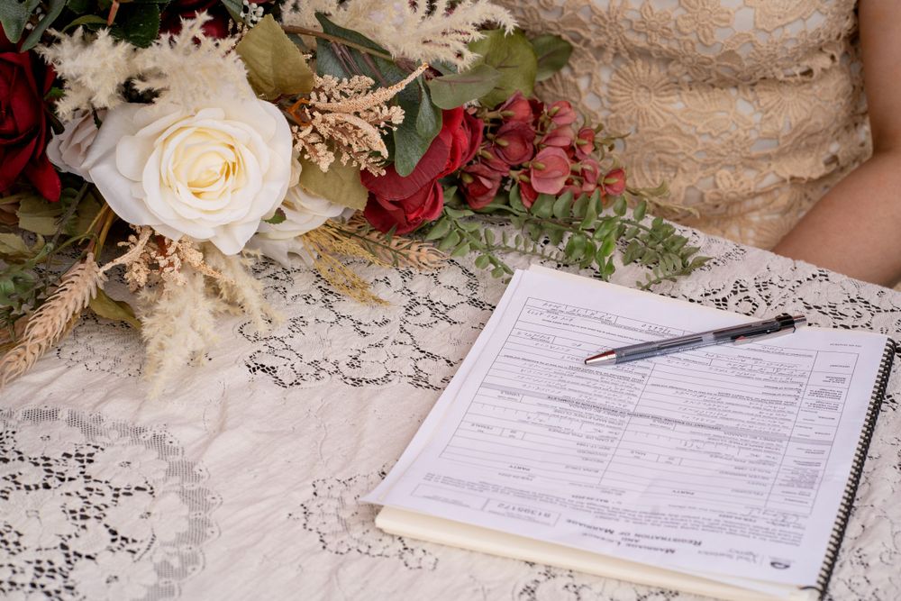 Marriage license form with pen lies next to elegant white and red floral bouquet on lace tablecloth.