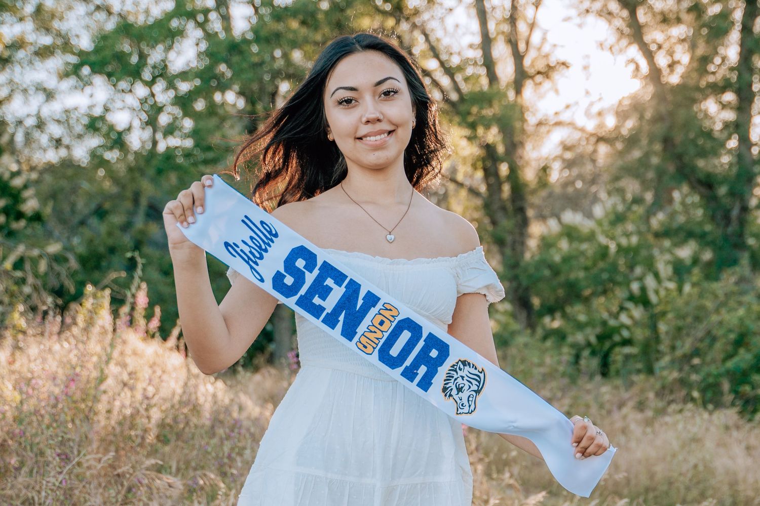 Woman in white off-shoulder top wearing senior sash outdoors during golden hour.