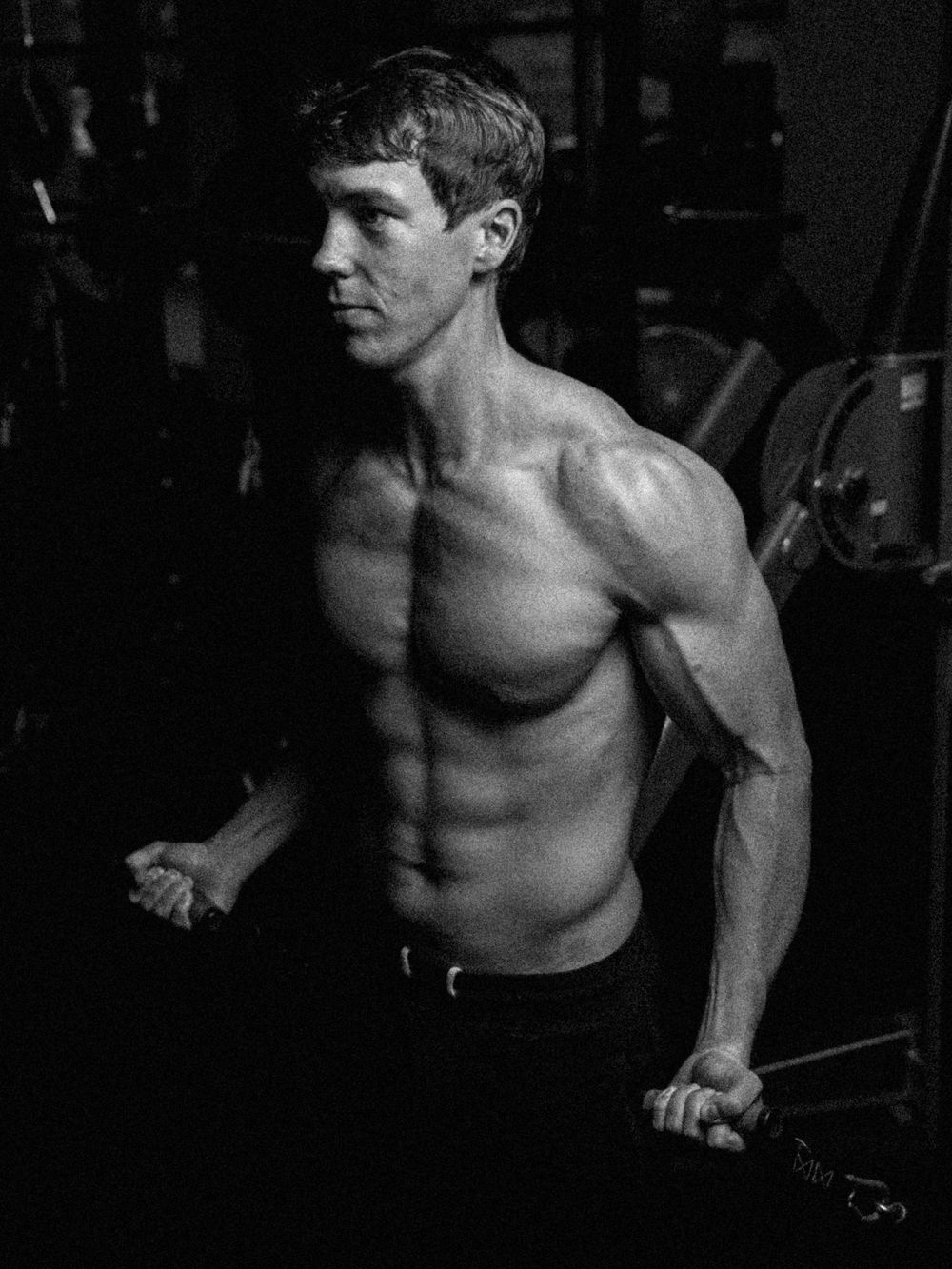 Black and white photograph of a muscular shirtless athlete performing a workout on dip bars in a dark gym setting.