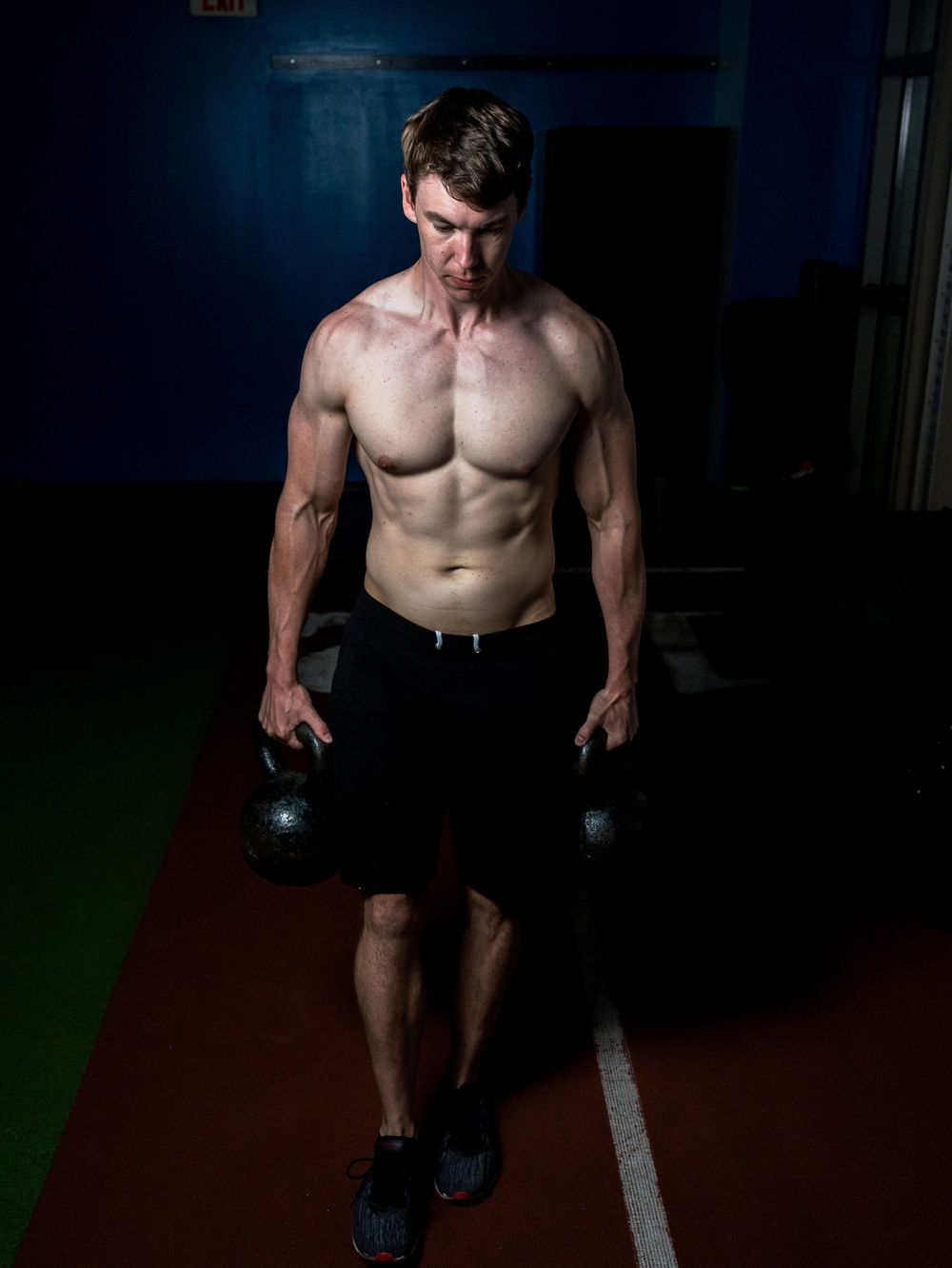 Shirtless athletic person holding kettlebells during an intense workout session in a dimly lit gym space.