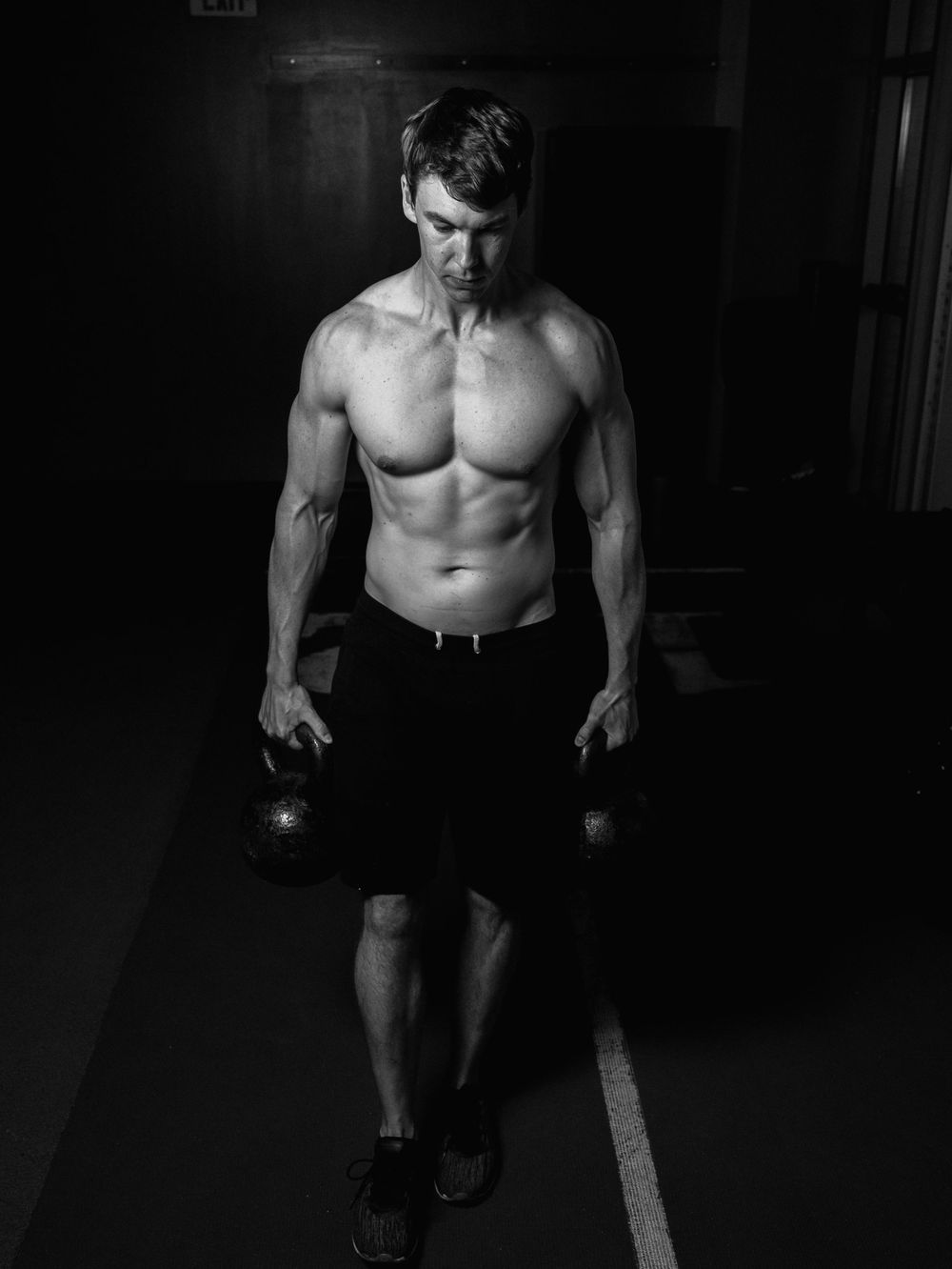 Athletic figure holding kettlebells while working out in a dark gym wearing black shorts in dramatic lighting.