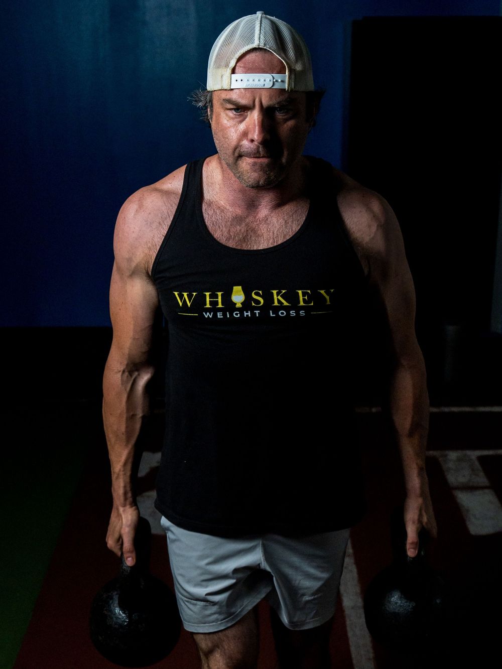 Athletic person in a black Whiskey tank top and baseball cap holding dumbbells in a dimly lit gym setting.