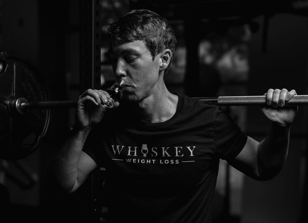 Person in dark clothing performs a barbell squat workout in dim lighting while wearing a Whiskey logo t-shirt.