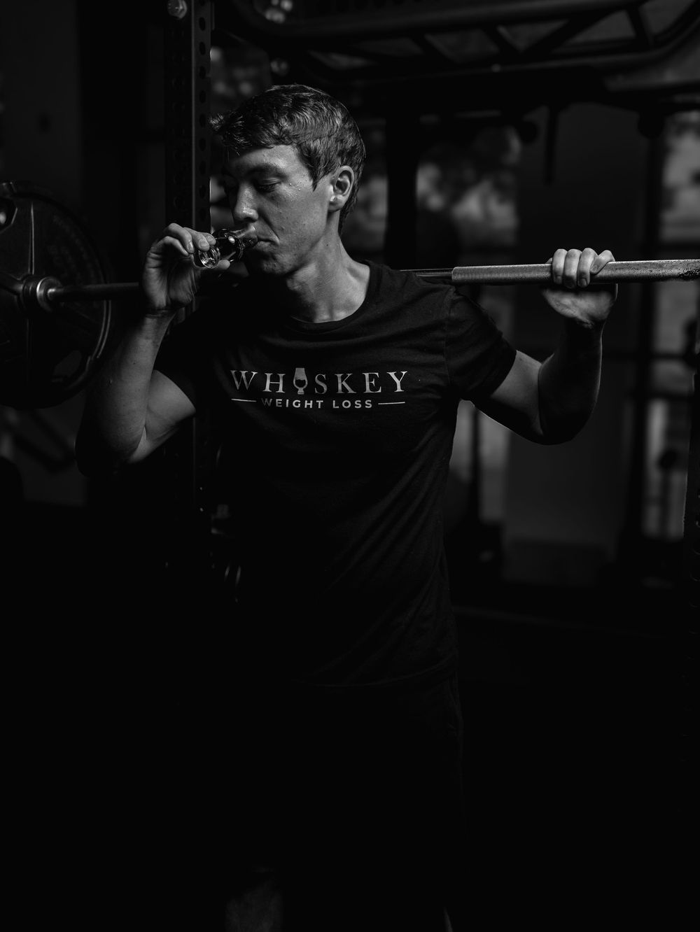 Black and white photo of an athlete in a Whiskey t-shirt performing a front squat with a barbell in a dark gym.