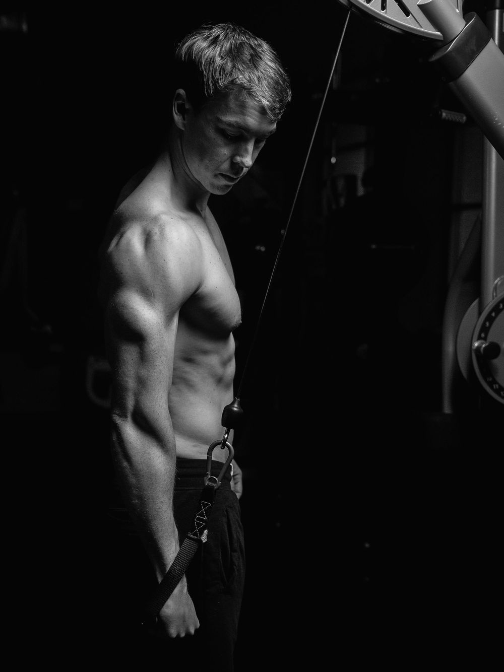 Black and white athletic physique photo showing muscular torso and arm against dark background in dramatic lighting.