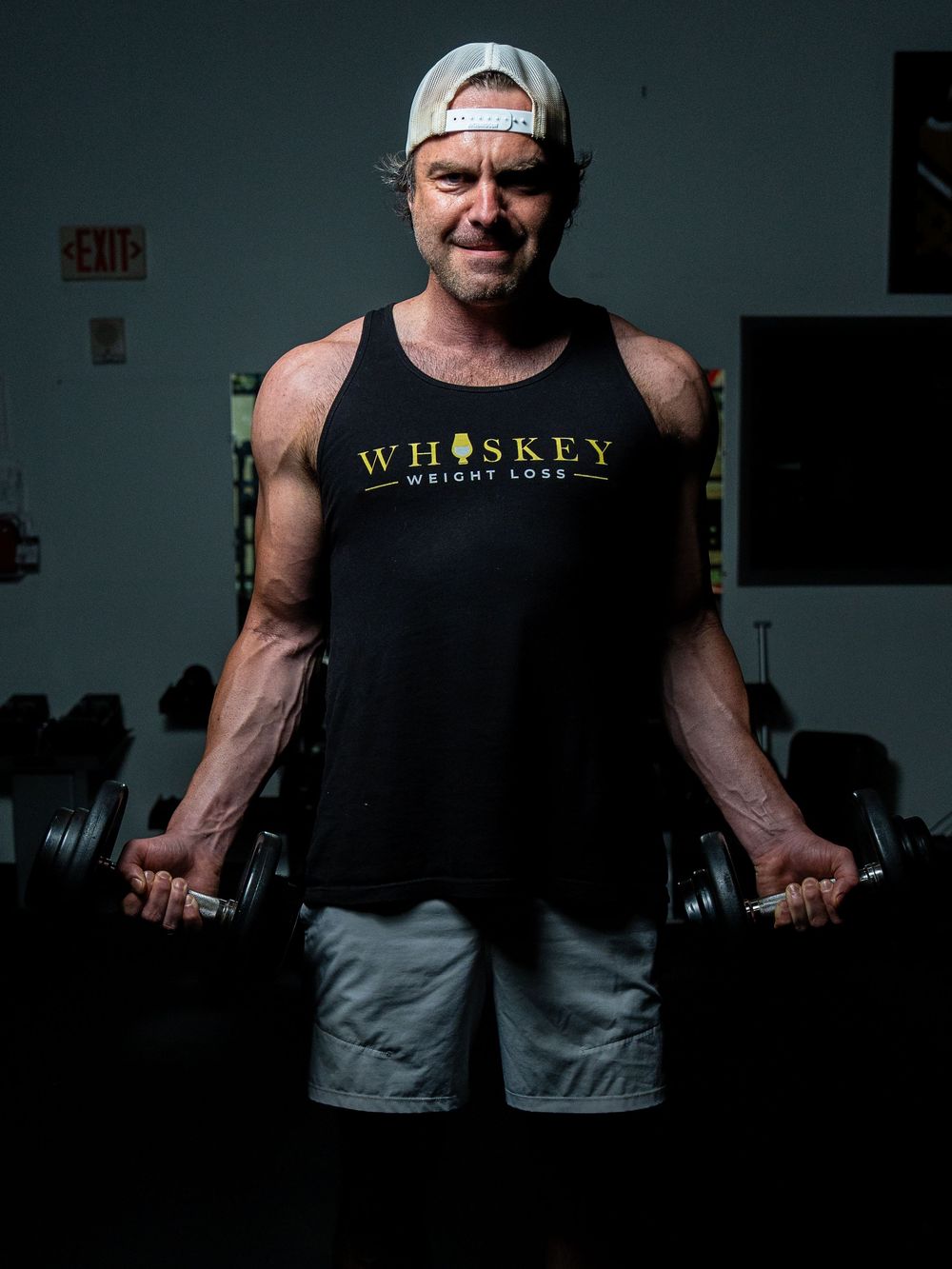 Person in a dark tank top performs bicep curls with dumbbells in a dimly lit gym setting during workout session.