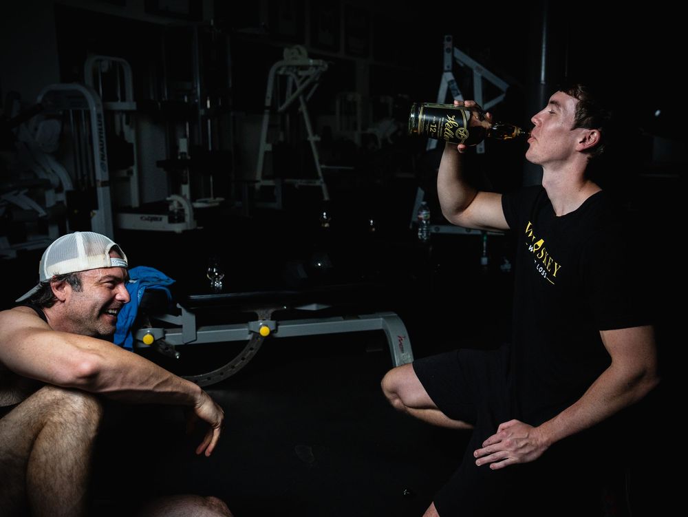 Two athletes take a break in a dark gym, with one person drinking from a water bottle while the other sits nearby.