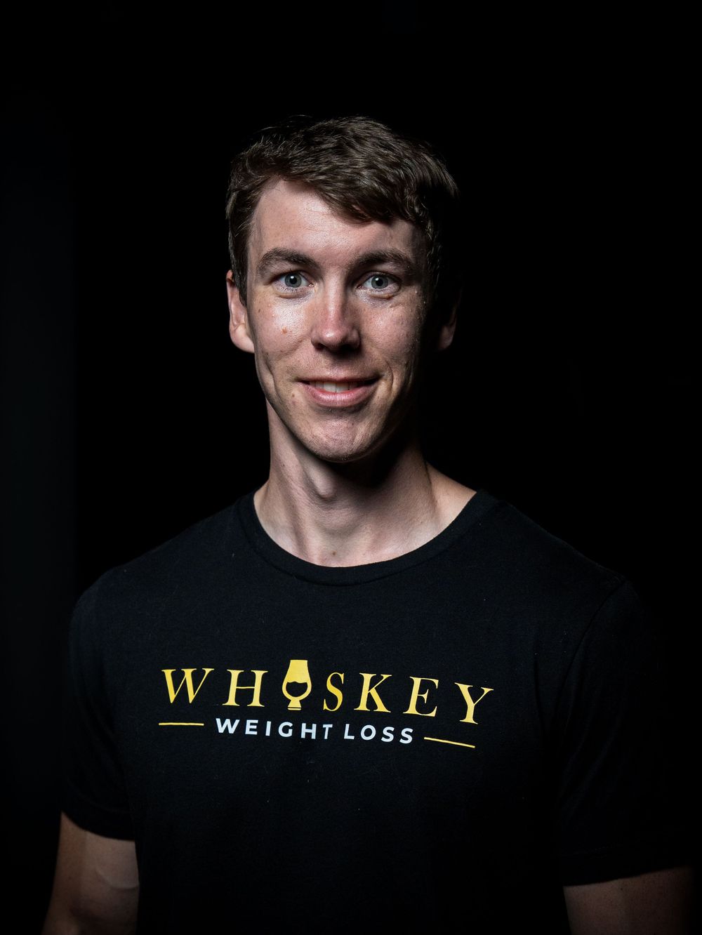 Portrait of someone wearing a black t-shirt with gold 'Whiskey Weight Loss' text against a dark background.