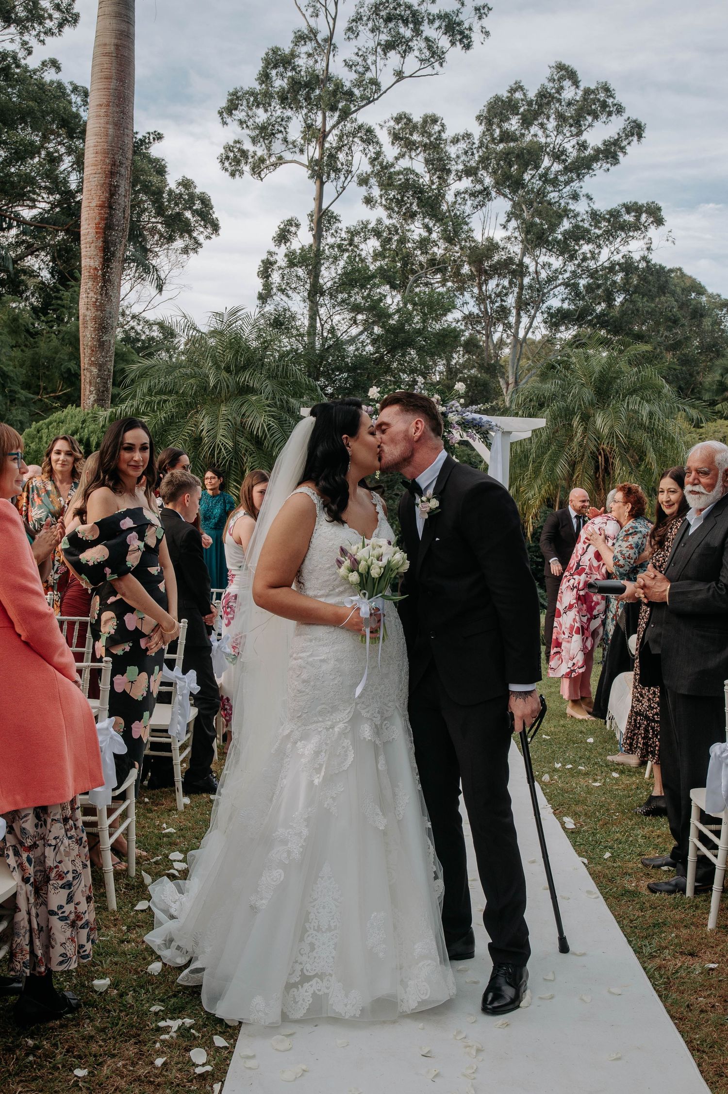 A couple shares a tender kiss at their outdoor garden wedding ceremony surrounded by guests on white chairs and palm trees.