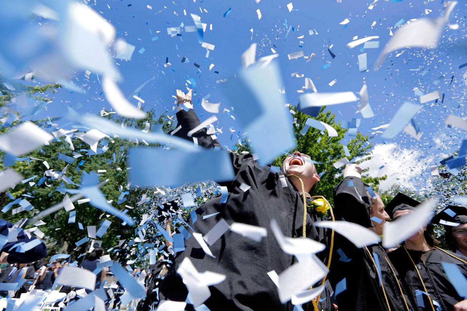 University graduates celebrate by throwing their cap while surrounded by blue and white confetti.