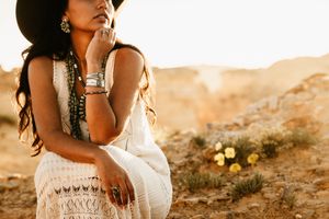 Boho-styled woman in lace dress and silver jewelry sits in desert landscape during golden hour.